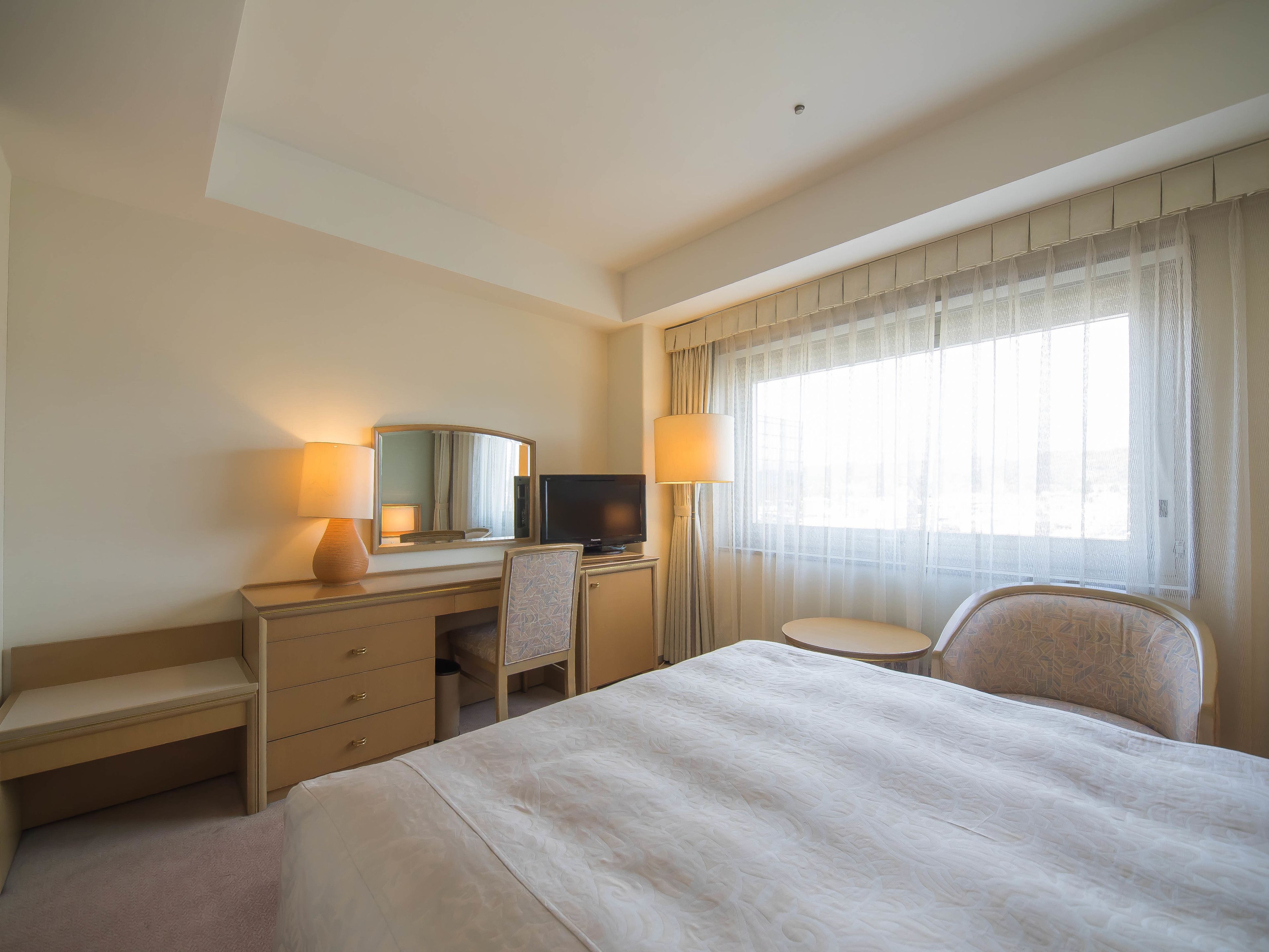 A hotel room with a bed, a desk with a chair, a television, and a window with sheer curtains.