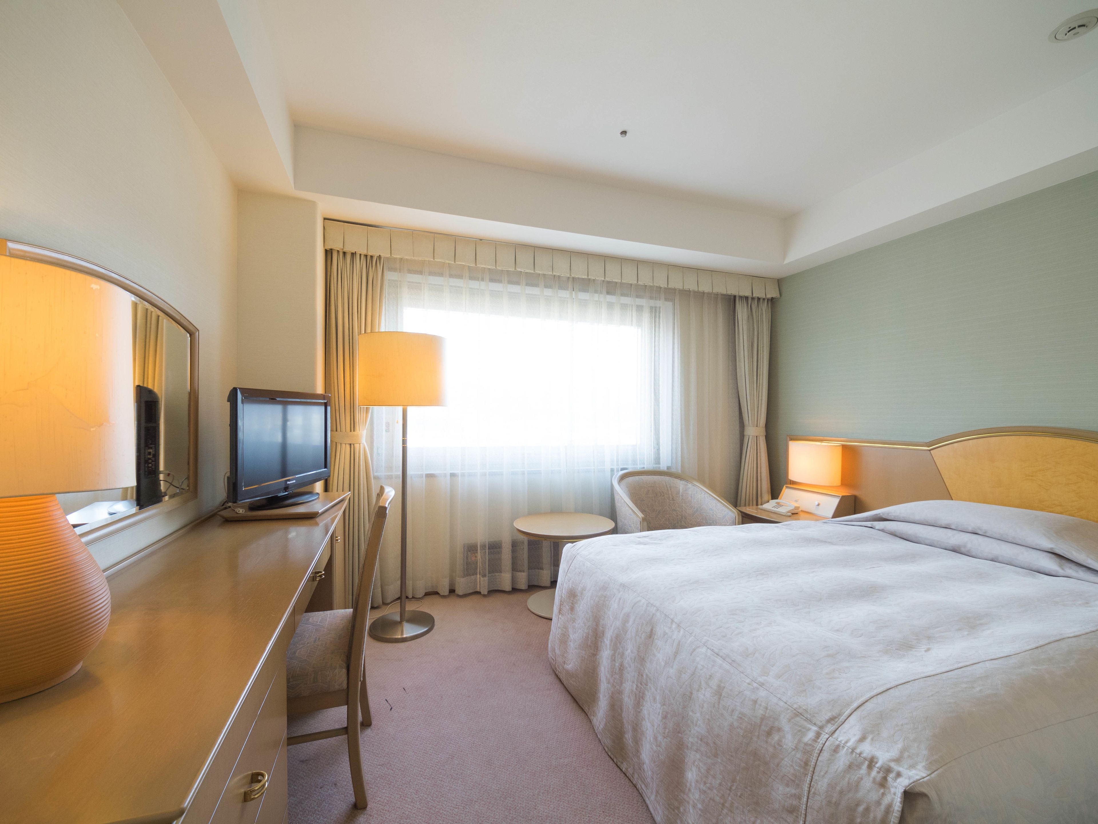 A hotel room with a bed, a desk with a television, a chair, and a lamp.