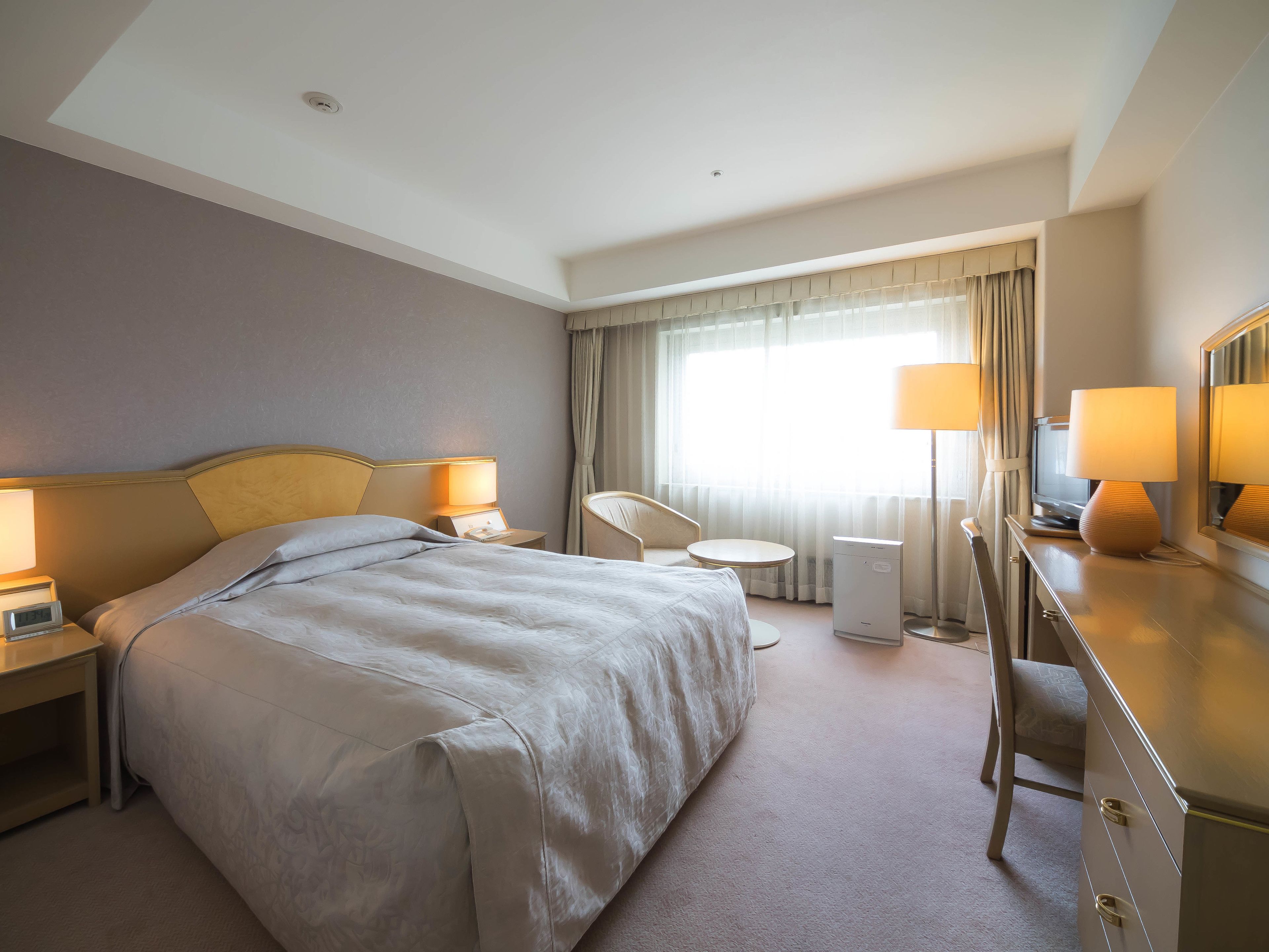 Habitación de hotel con cama, mesitas de noche, escritorio, silla, espejo y ventana con cortinas.