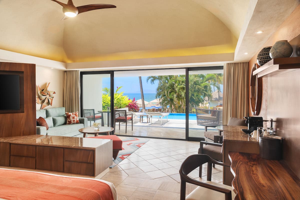 Habitación de hotel con un amplio ventanal corredera que da a una zona de piscina, un sofá, una mesa de comedor y una cocina.