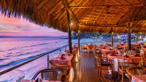Un restaurante con mesas y sillas, techo de paja y vista al océano.