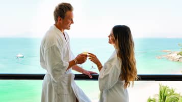 A couple in white robes by a window overlooking a beach with a boat in the distance.
