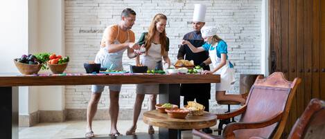 A group of people preparing food at a wooden table with bowls of fruits and vegetables, in a room with a brick wall and wooden door.