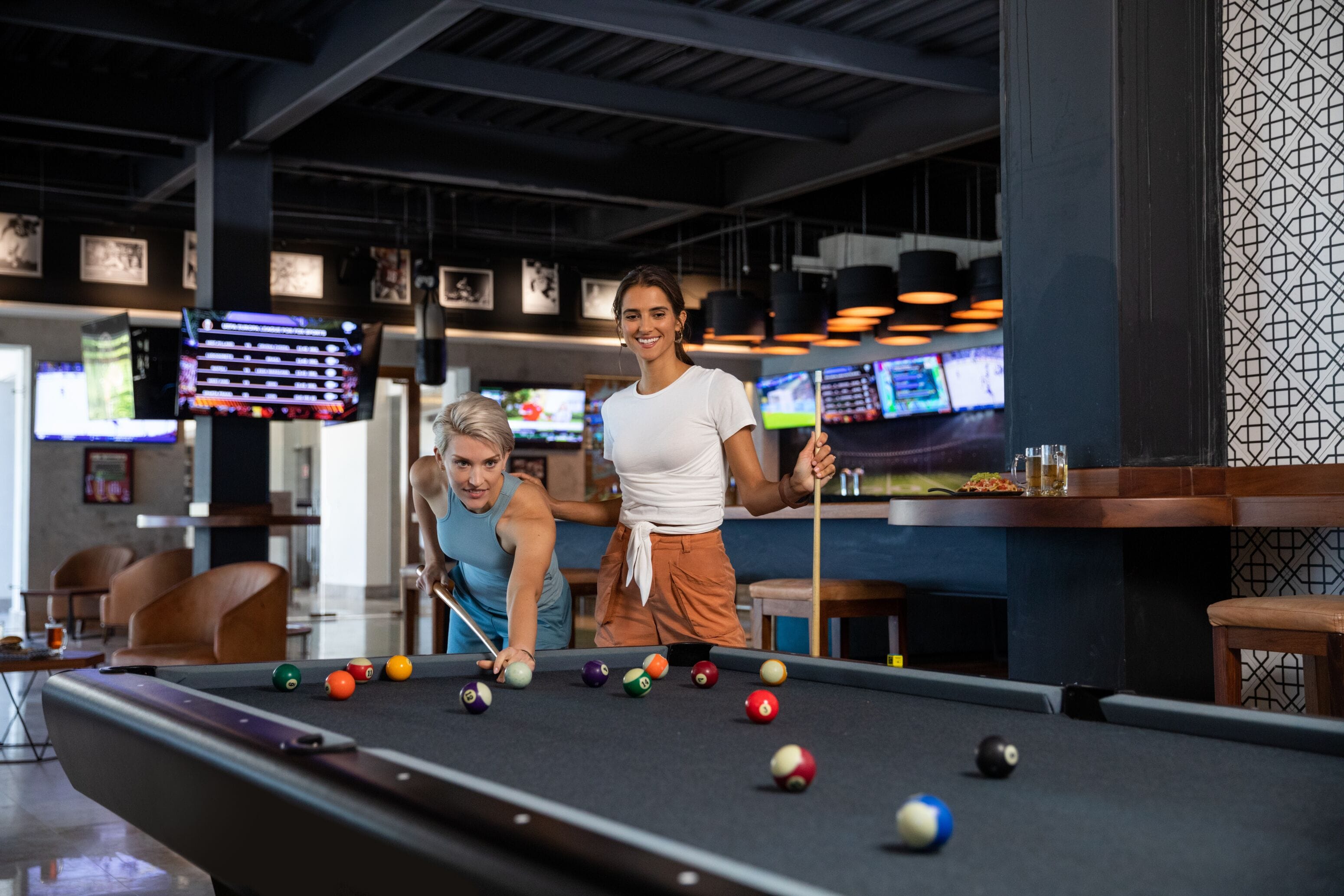 Une table de billard dans un décor intérieur moderne, avec plusieurs écrans diffusant des événements sportifs en arrière-plan.