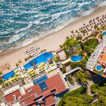 An aerial view of a resort with a beach, a swimming pool, and a large building with multiple balconies.