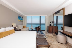 A hotel room with a large bed, a desk, a chair, a TV, and a view of the sea.