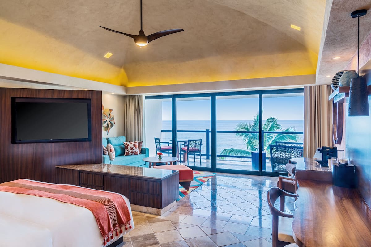 Habitación de hotel con una cama grande, zona de estar con vista al océano, comedor y televisor montado en la pared.