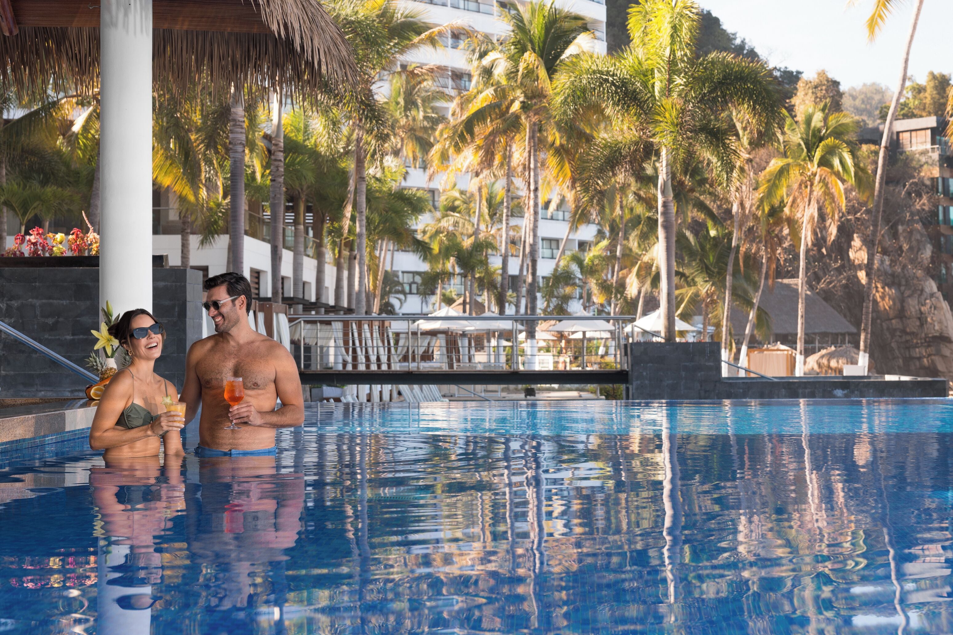 A swimming pool with a couple standing in it, surrounded by palm trees and a building in the background.