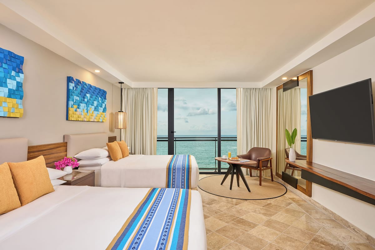 Habitación de hotel con dos camas, un escritorio, una silla, un televisor y un ventanal con vista al mar.