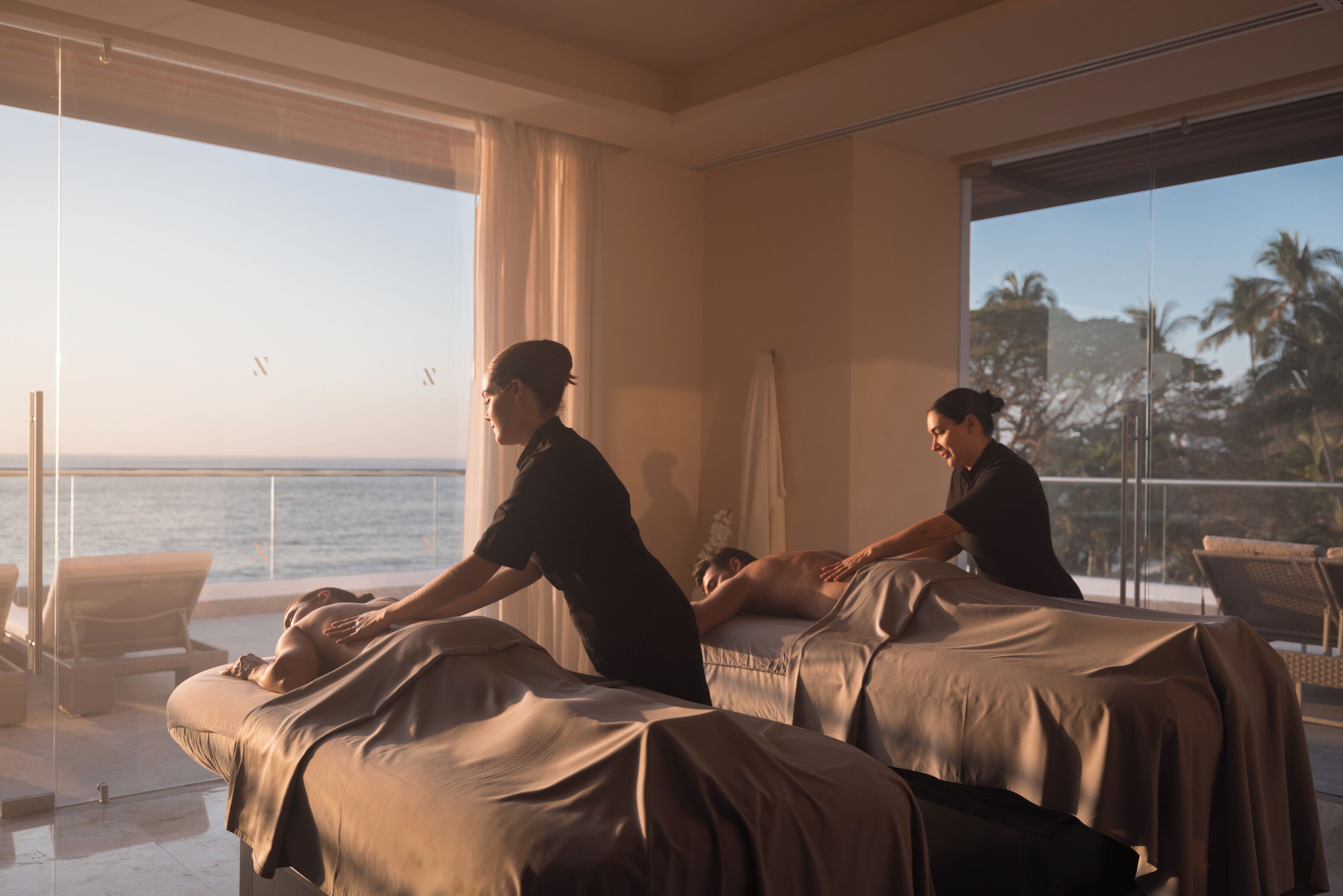 Un cuarto de terapia de masajes con dos camillas, ventanas amplias con vista al océano y un balcón con sillas.