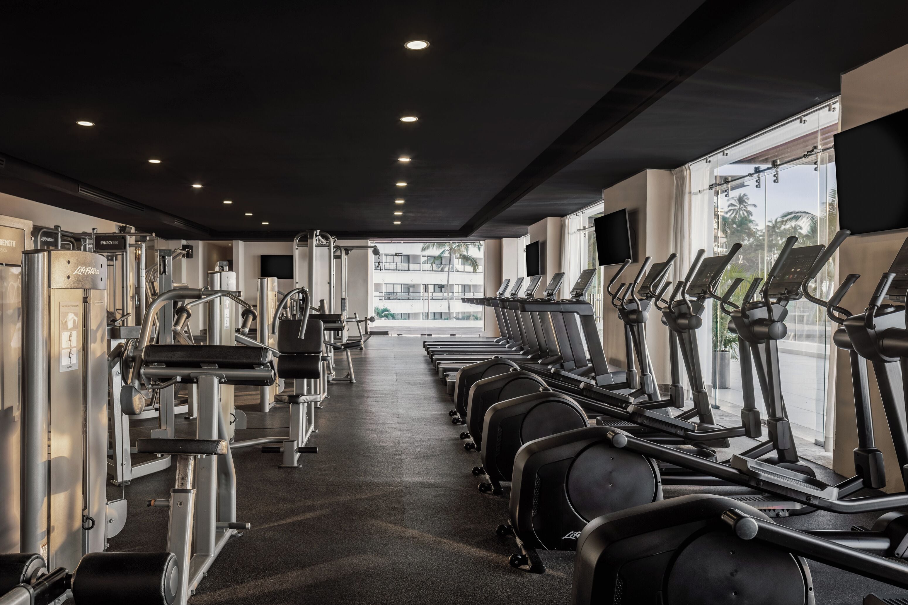 Ein Fitnessstudio mit Laufbändern, Krafttrainingsgeräten und einem großen Fenster mit Blick auf Palmen.