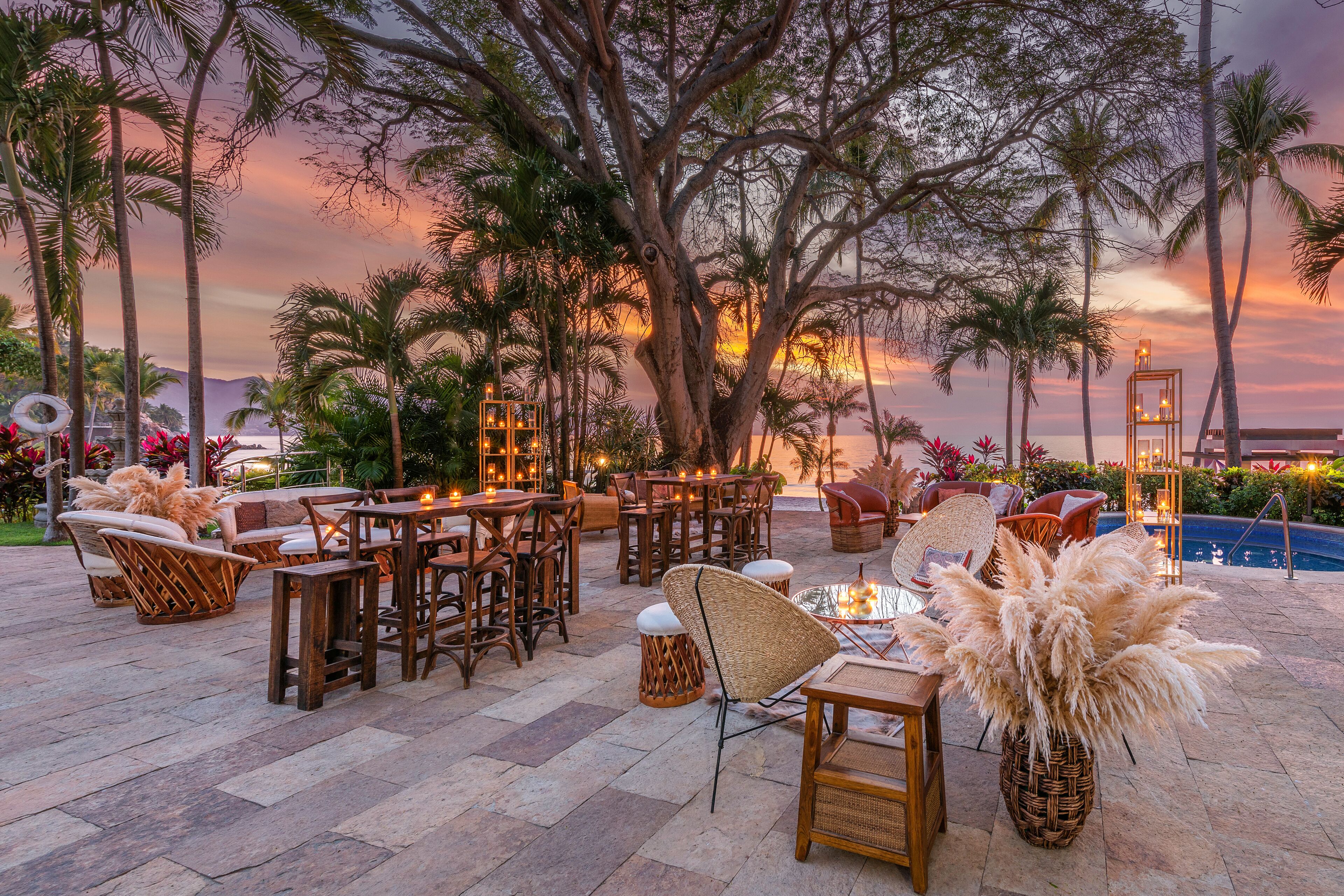 Un área de estar al aire libre con mobiliario de mimbre y velas, rodeada de palmeras y vista al atardecer.