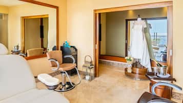 Quarto de hotel com uma cama grande, área de estar com uma poltrona e uma pequena mesa, toucador com espelho e vista para o mar através de uma porta de vidro.