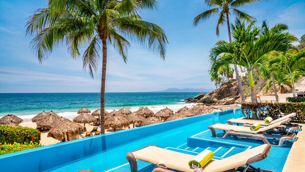 Zona junto a la piscina con sillones de descanso, vista despejada del océano y sombrillas de paja.