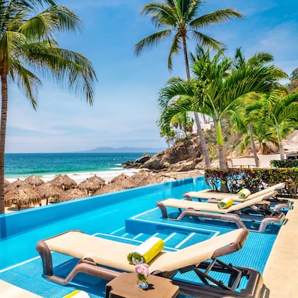 A poolside area with lounge chairs, a clear view of the ocean, and thatched umbrellas.