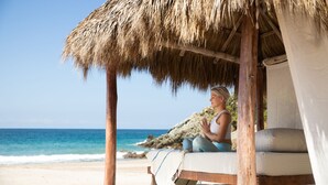 Una cabaña en la playa con techo de paja, un banco acolchado y vista al océano.
