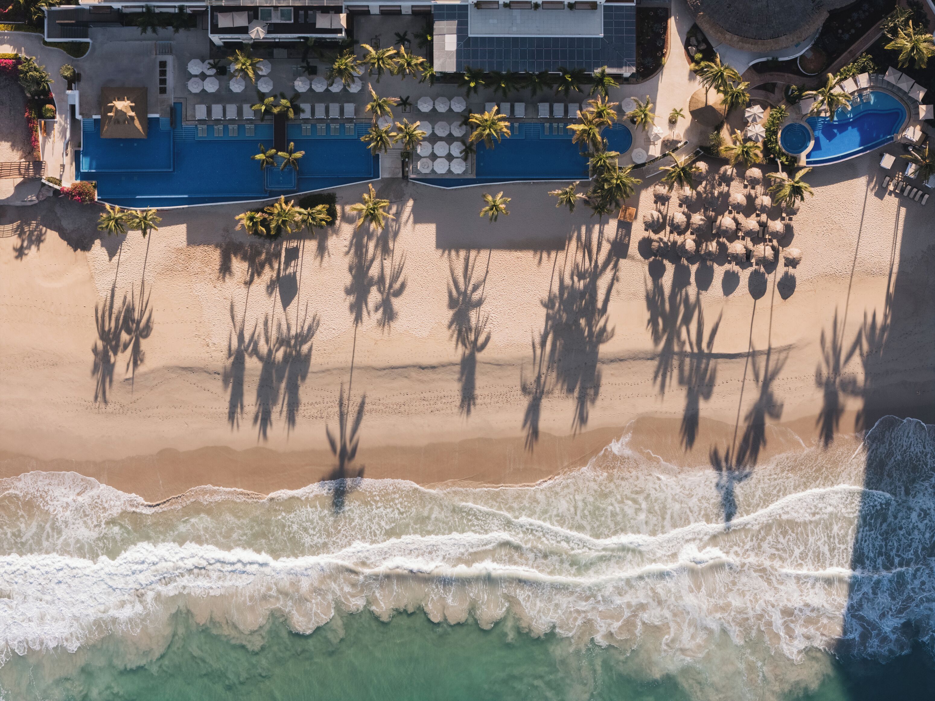 An aerial view of a beachfront hotel with a swimming pool, sun loungers, and palm trees.