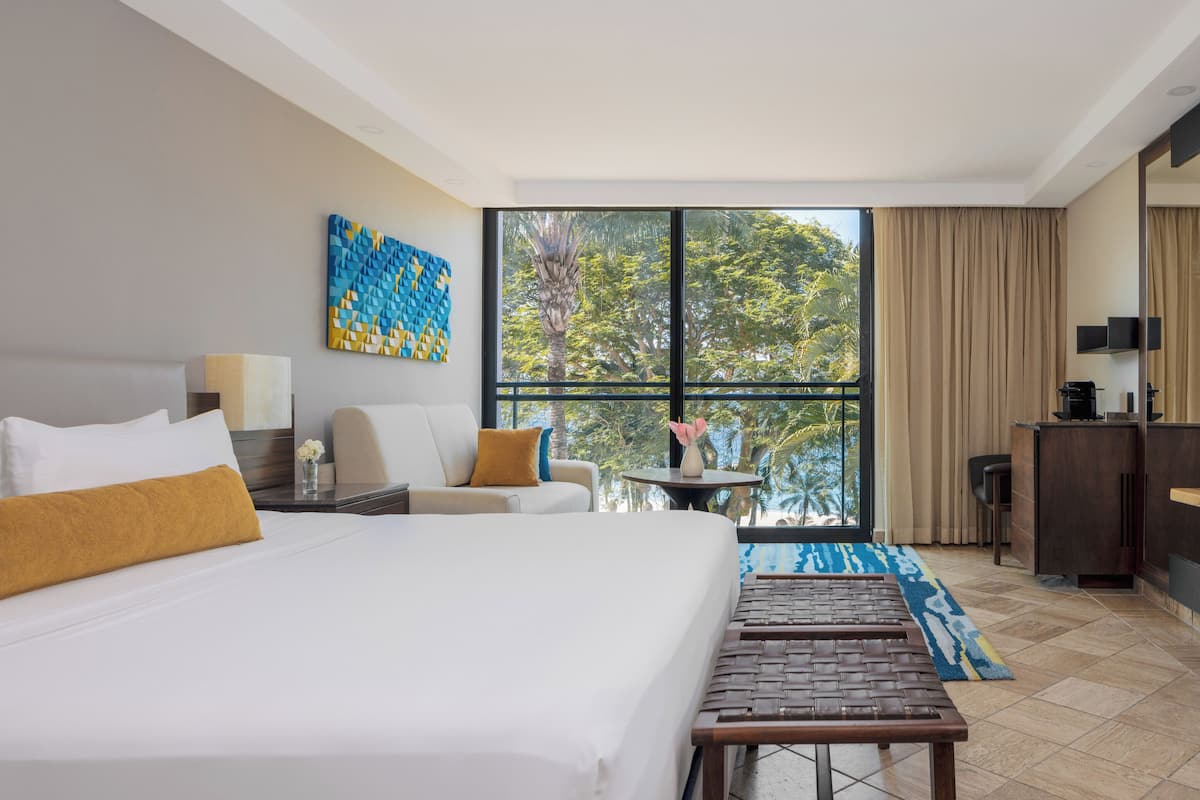 Habitación de hotel con una cama grande, un escritorio, una silla, una mesita con un jarrón y vistas a árboles a través de un ventanal amplio.