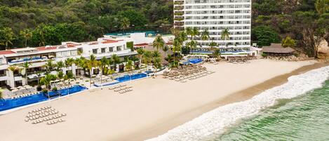 Un hotel frente a la playa con varias piscinas, una playa con sillas de descanso y un edificio grande con balcones.
