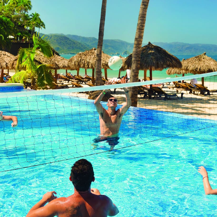 A pool area with a volleyball net, palm trees, and a beach in the background.