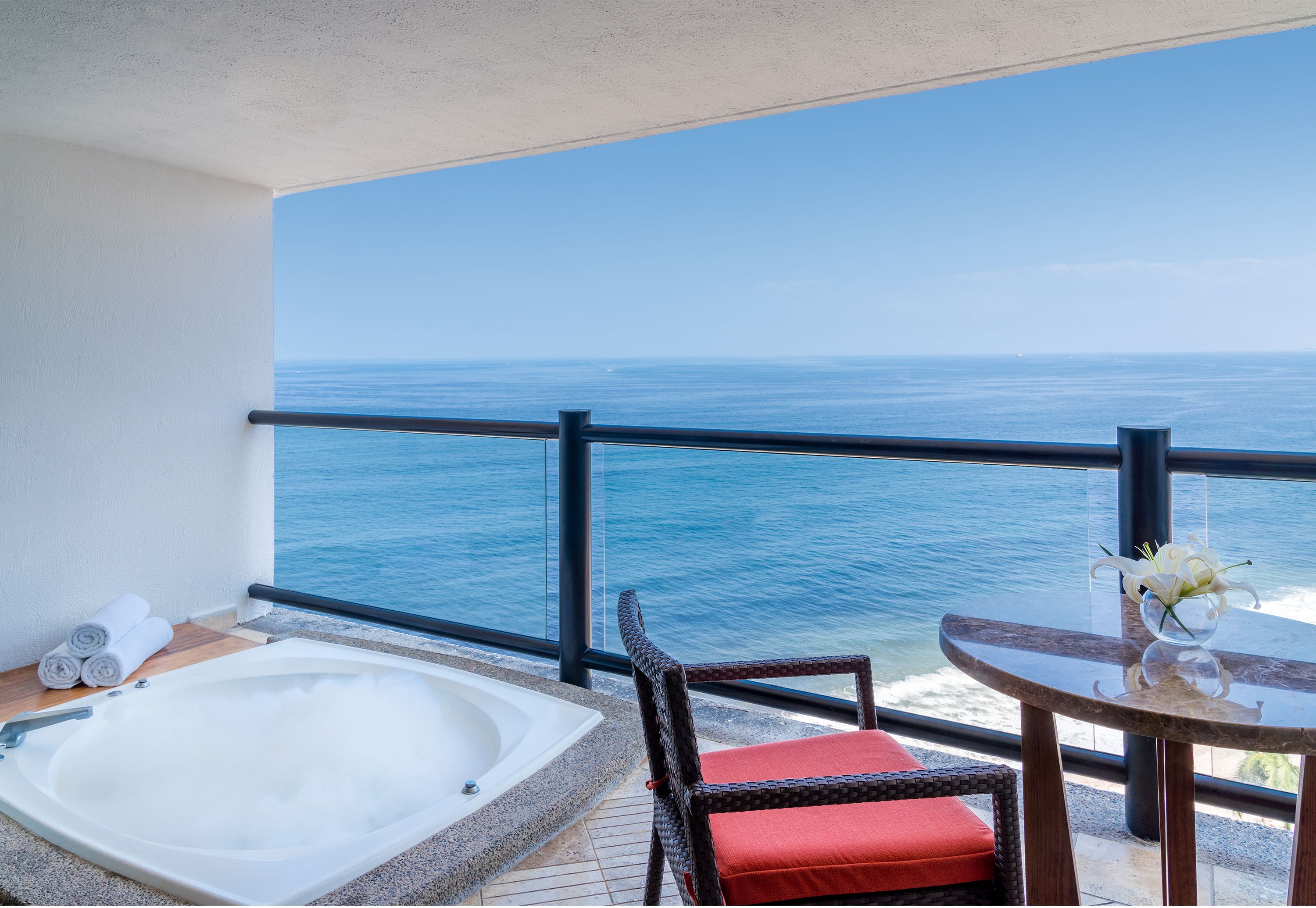 Un balcón con vista al océano, un jacuzzi, una silla de mimbre y una mesa con un jarrón de flores.