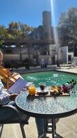 Una persona sentada a una mesa con una bandeja de comida y bebidas, con vista a una piscina y una clara visión de la arquitectura circundante.