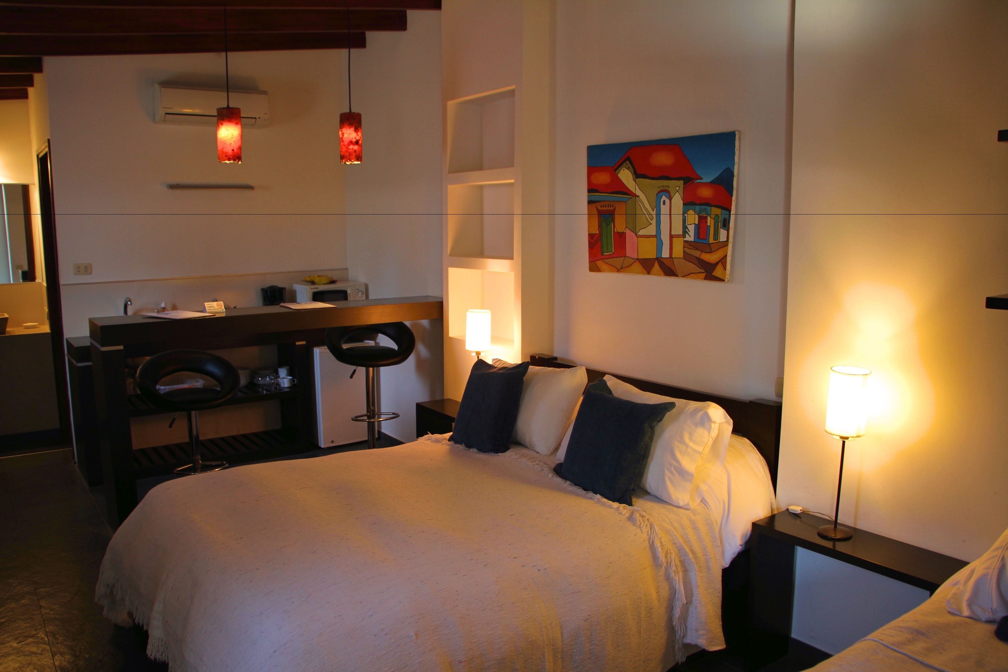 A hotel room with a bed, a desk, a chair, a painting, and a nightstand with a lamp.