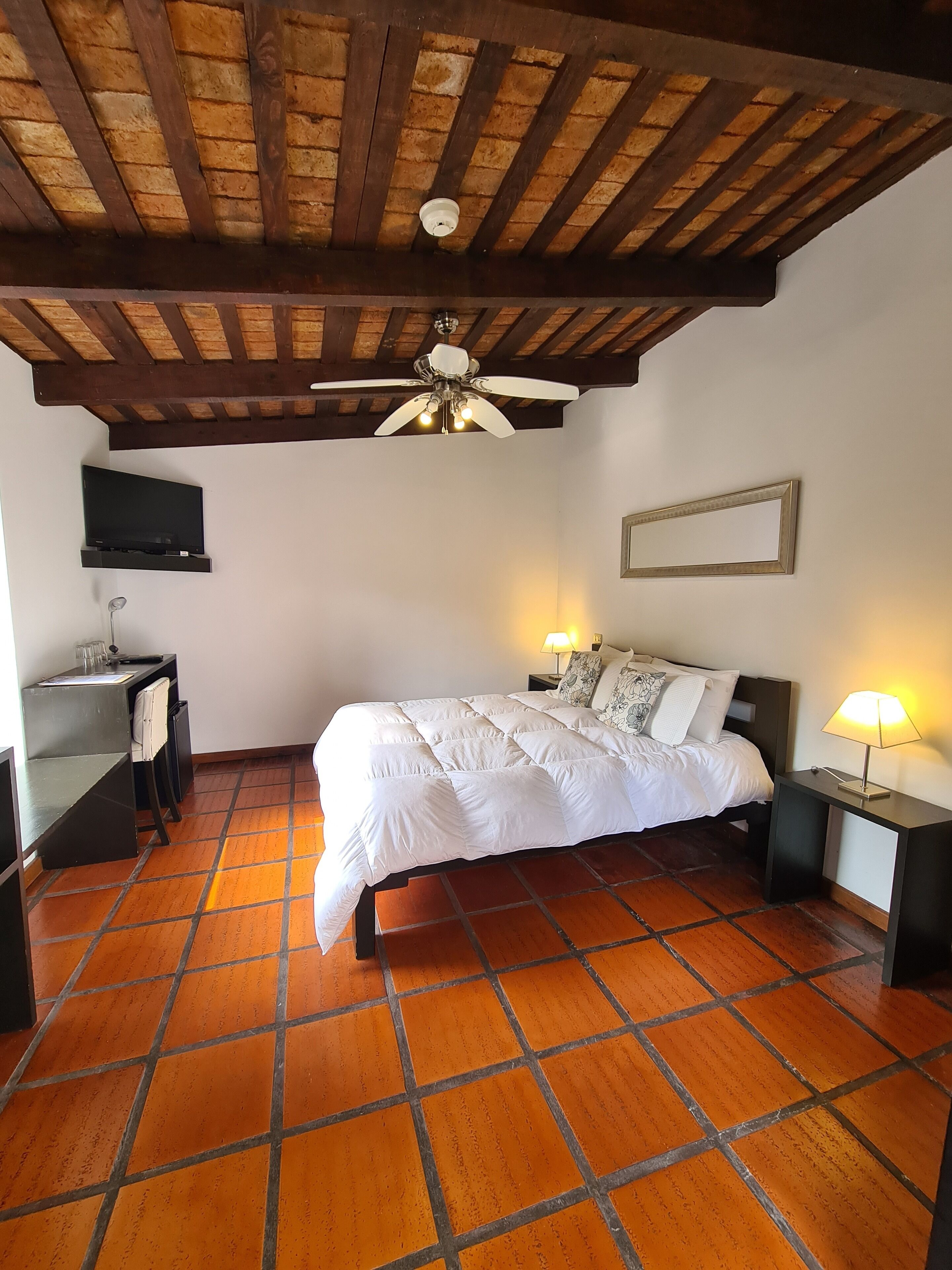 A bedroom with a bed, a ceiling fan, a TV, a desk, and a nightstand with a lamp.