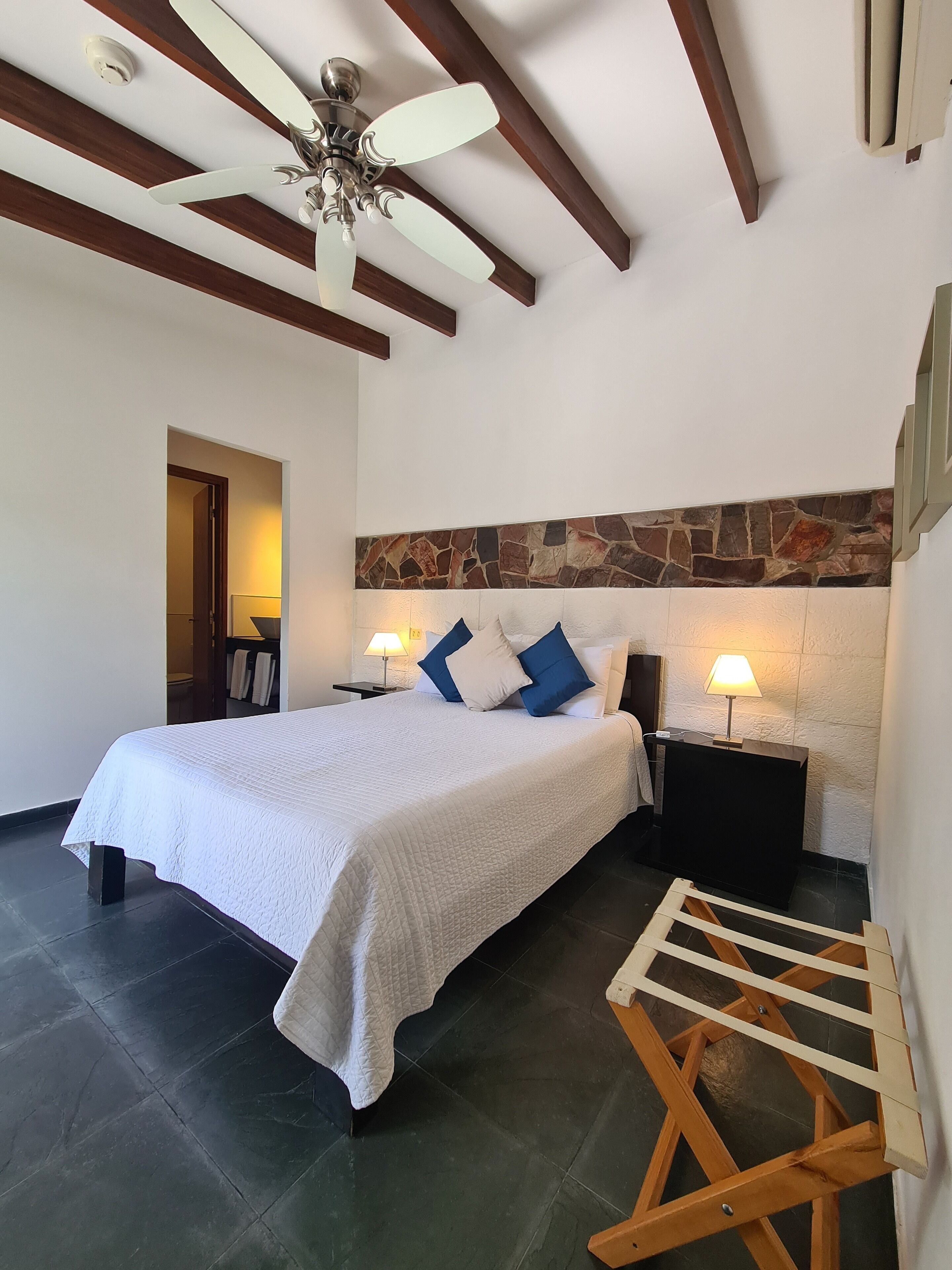 A bedroom with a bed, two bedside tables with lamps, a ceiling fan, a wooden ladder, and a stone accent wall.