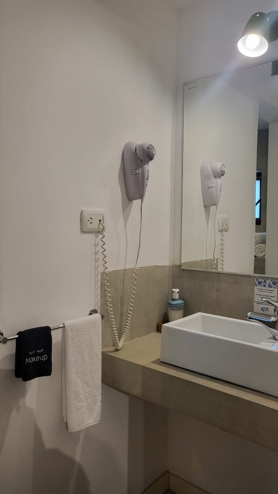 Un baño con un lavamanos blanco, un perchero con una toalla negra y dos secadores de pelo fijados a la pared.