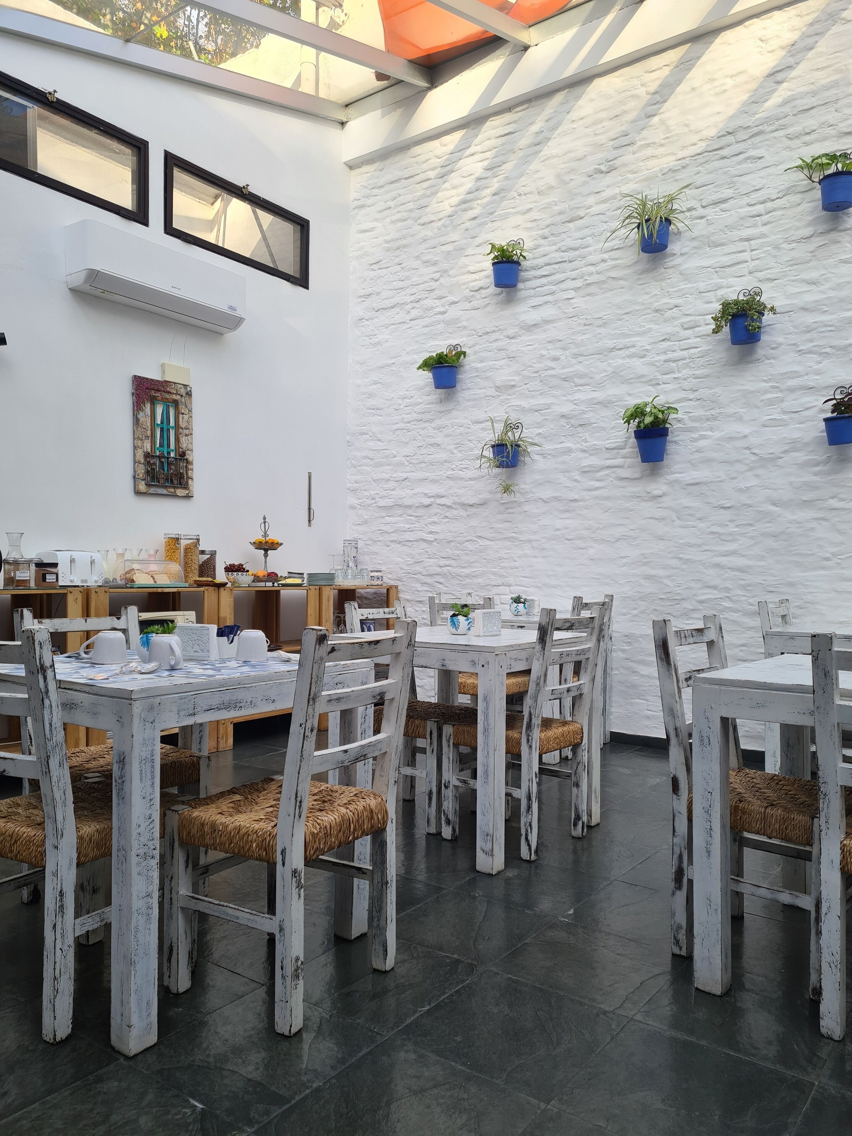 A dining area with white wooden tables and chairs, potted plants on the wall, and a stone wall backdrop.