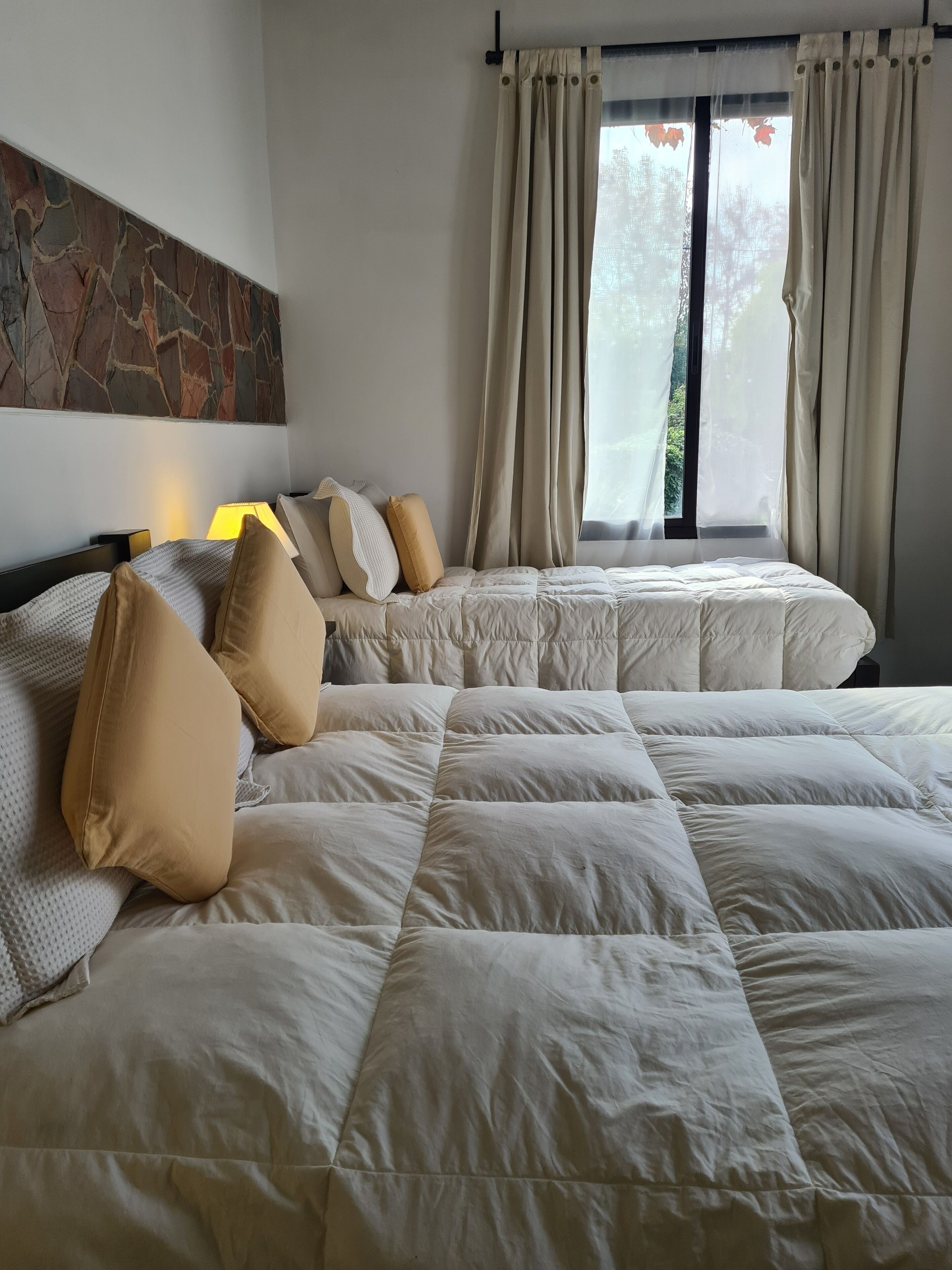 A hotel room with two beds, beige bedding, a stone accent wall, and a window with curtains.
