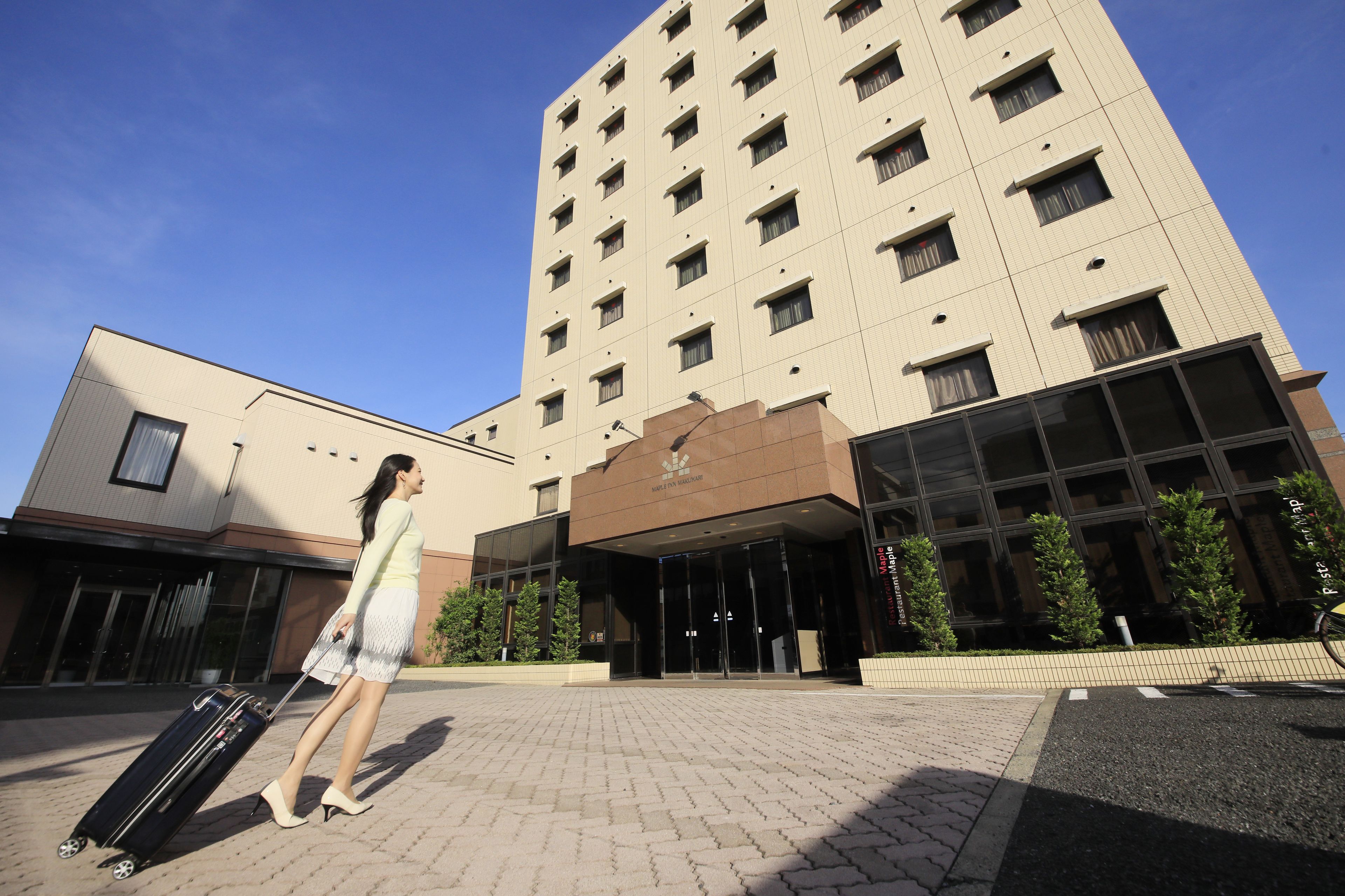 A modern hotel entrance with a glass facade, a brick-paved area, and a person with a suitcase.