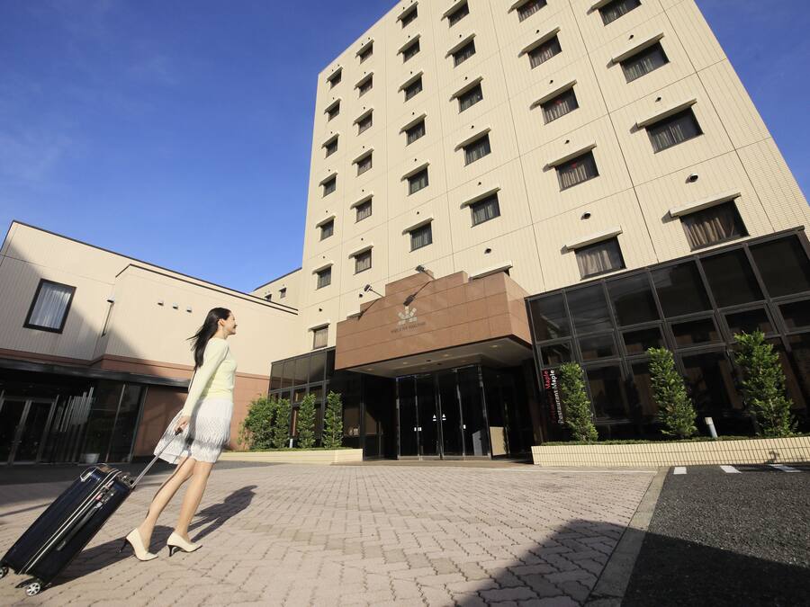 A modern hotel entrance with a glass facade, a brick-paved area, and a person with a suitcase.