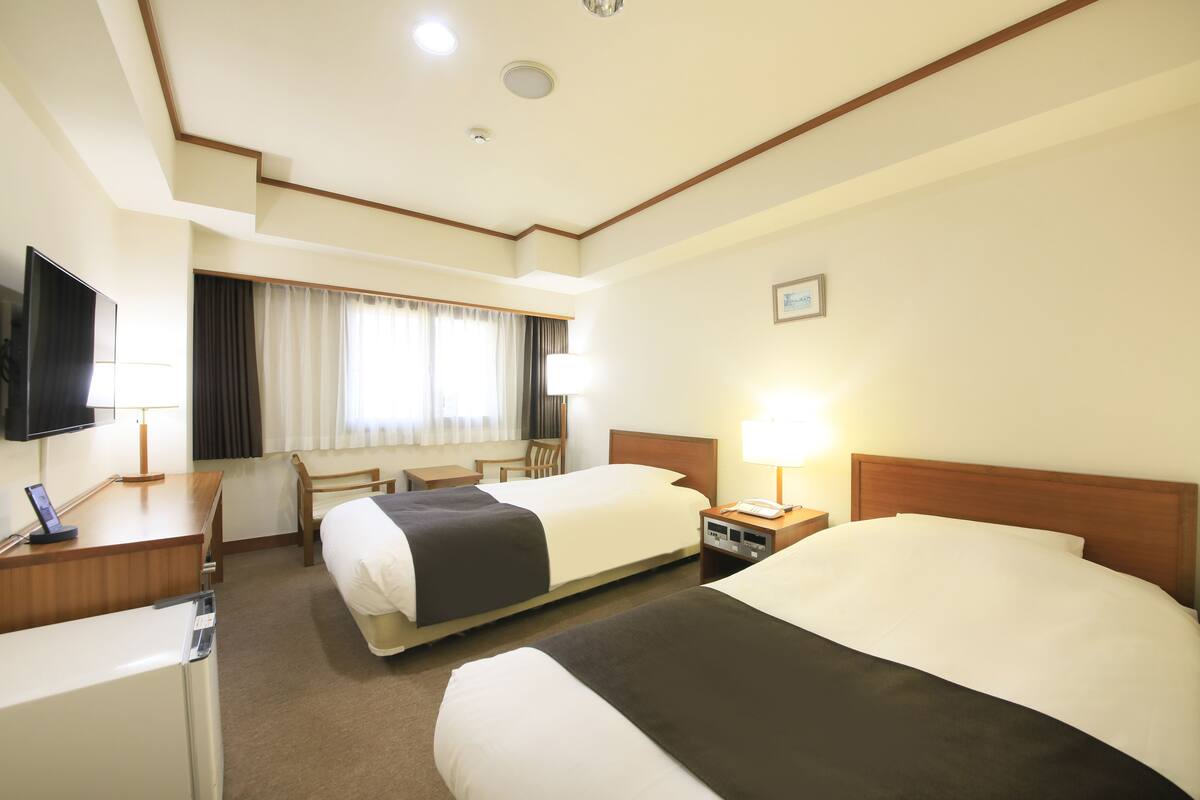 A hotel room with two beds, a desk with a TV, a chair, a lamp, and a window with curtains.
