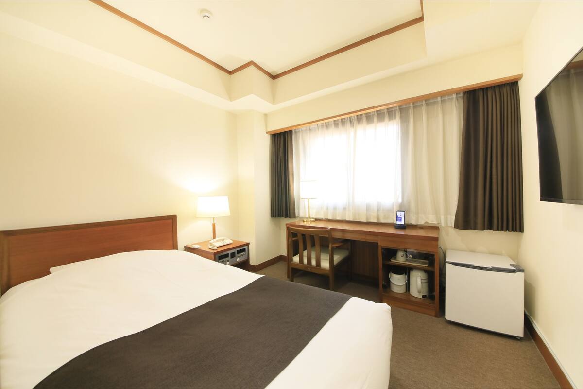 A hotel room with a large bed, a desk, a chair, a desk lamp, a phone, a mini-fridge, and a window with curtains.