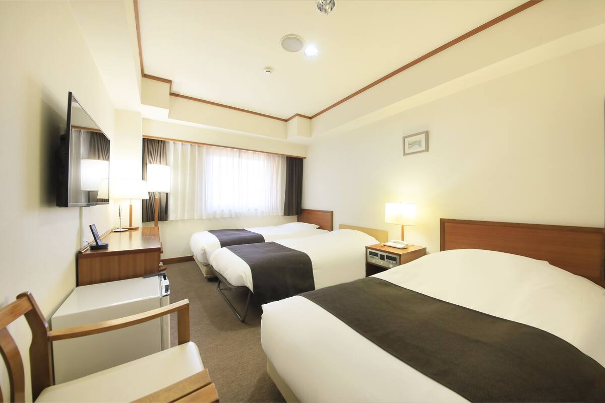 A hotel room with three beds, a desk, a chair, a TV, and a window with curtains.