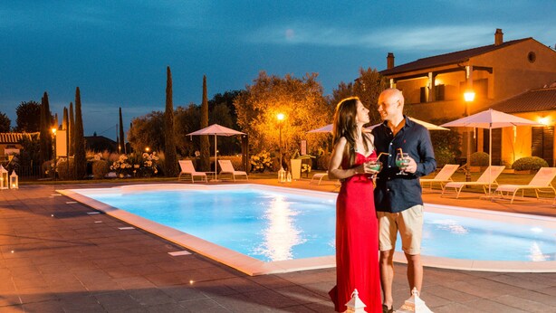 Una pareja de pie junto a una piscina con sillones y sombrillas al fondo.