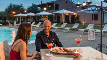 Una pareja cenando en una mesa al aire libre, con una piscina y sombrillas al fondo.