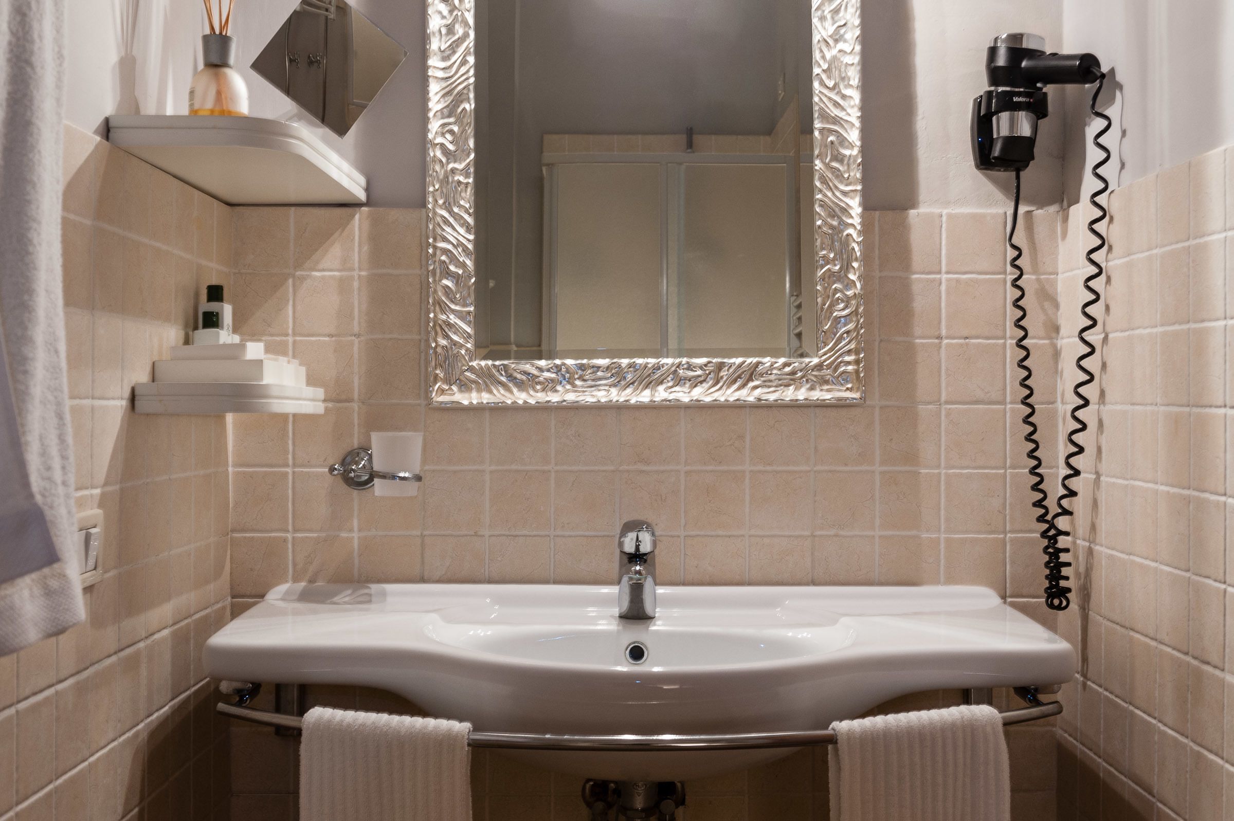 A bathroom with a white sink, a towel rack, a mirror with a decorative frame, and a hairdryer on the wall.