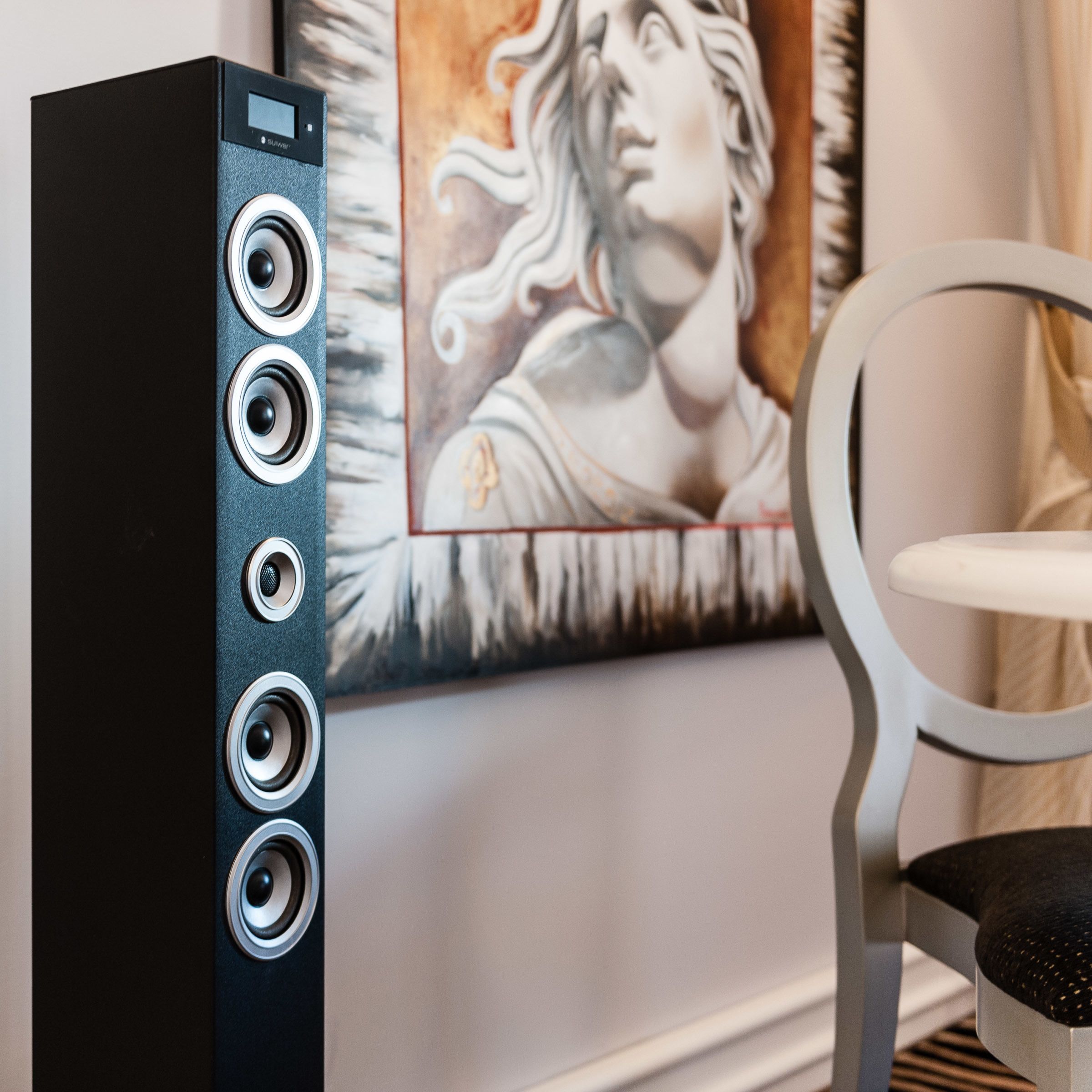 A black audio system with four speakers, a white chair, and a painting of a woman on the wall.