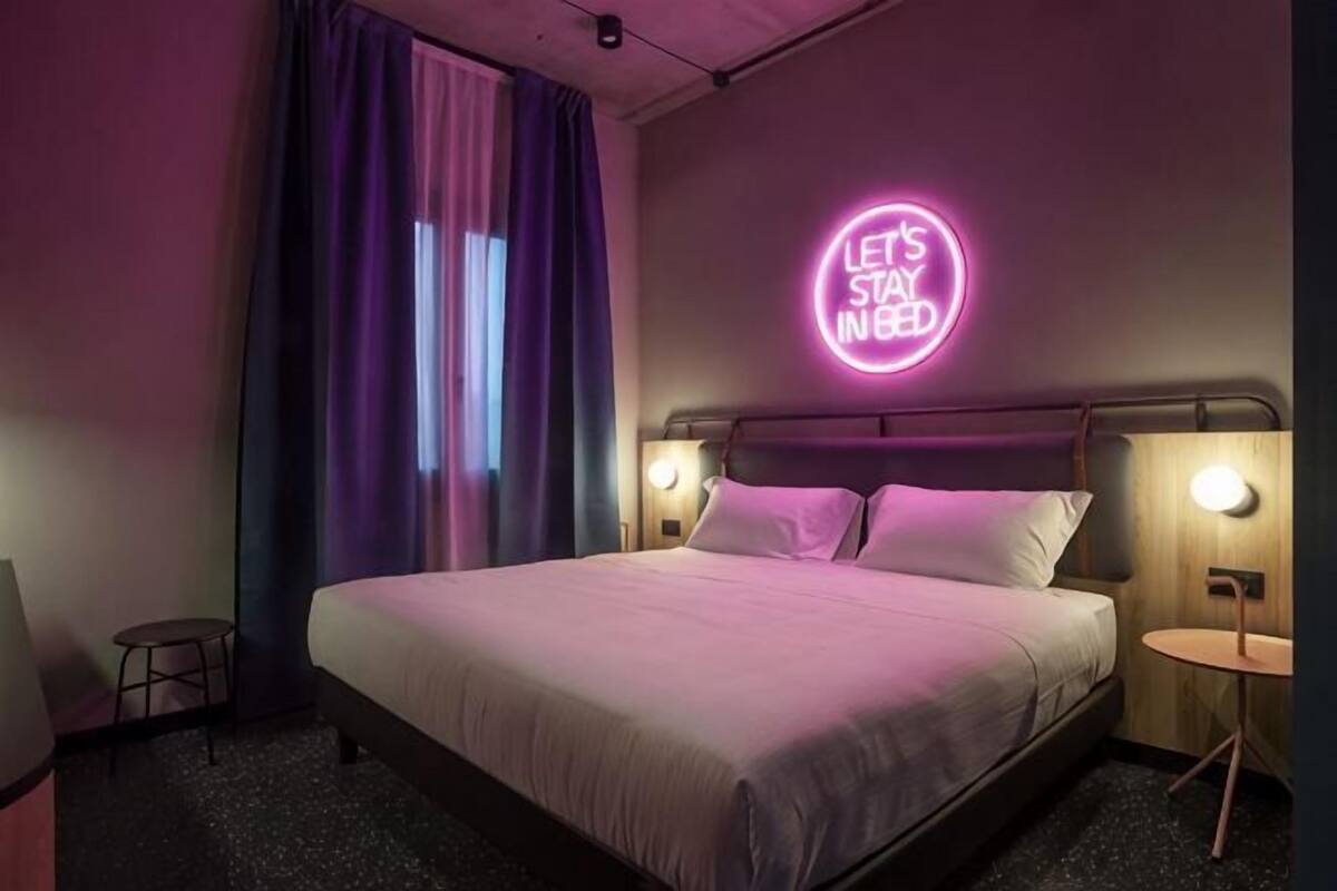 A hotel room with a bed, a neon sign, a window with curtains, and a small table.