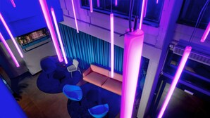 A modern interior with a seating area featuring round tables and chairs, a blue curtain backdrop, and neon lighting. - Stay Hotel (Brunello)