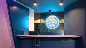 A reception desk with a neon sign reading "STAY HOTEL" and a glass partition. - Stay Hotel (Brunello)