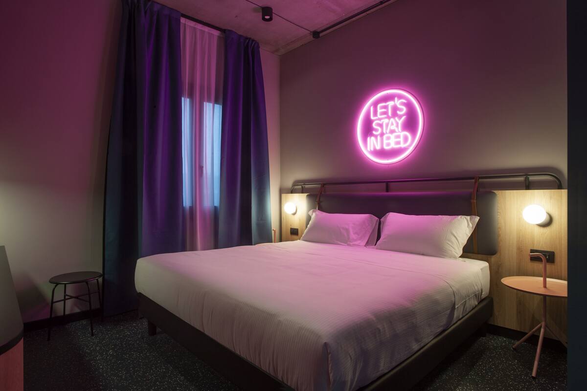 A modern hotel room with a bed, a neon sign on the wall, and a small table by the bed.