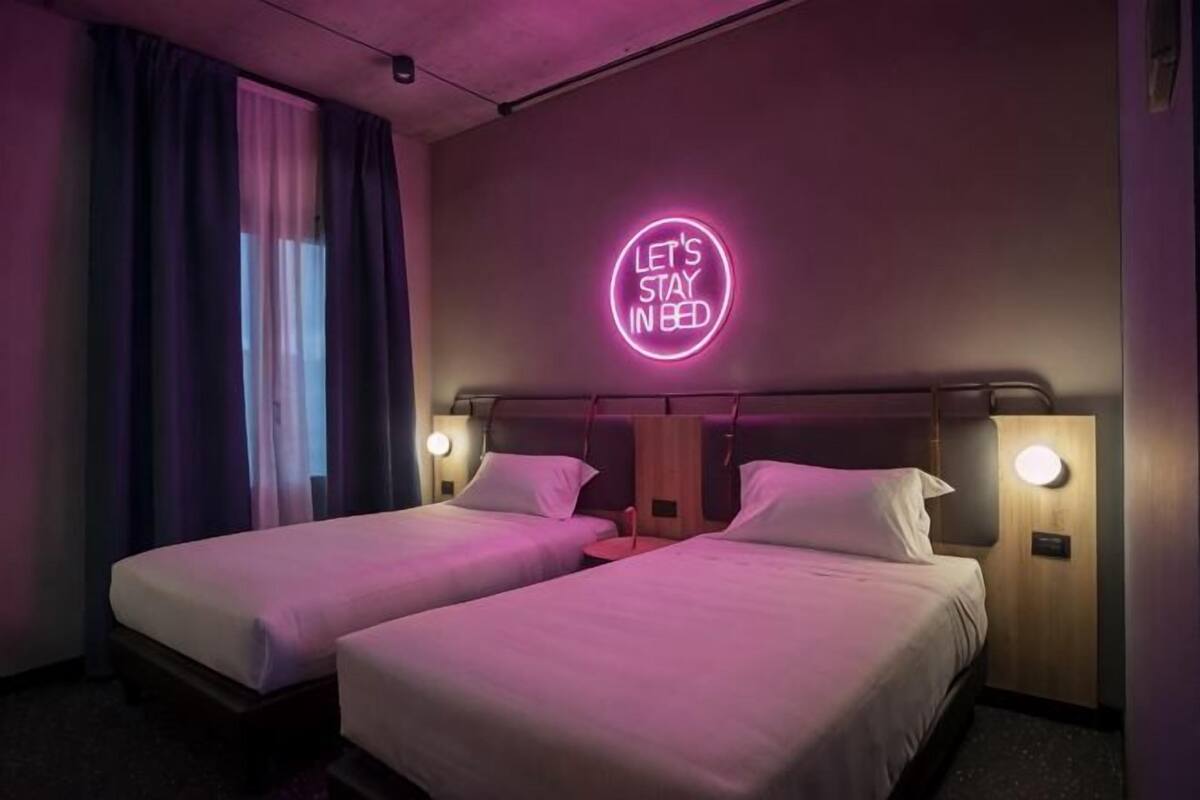 A hotel room with two beds, a neon sign on the wall, and a window with curtains.