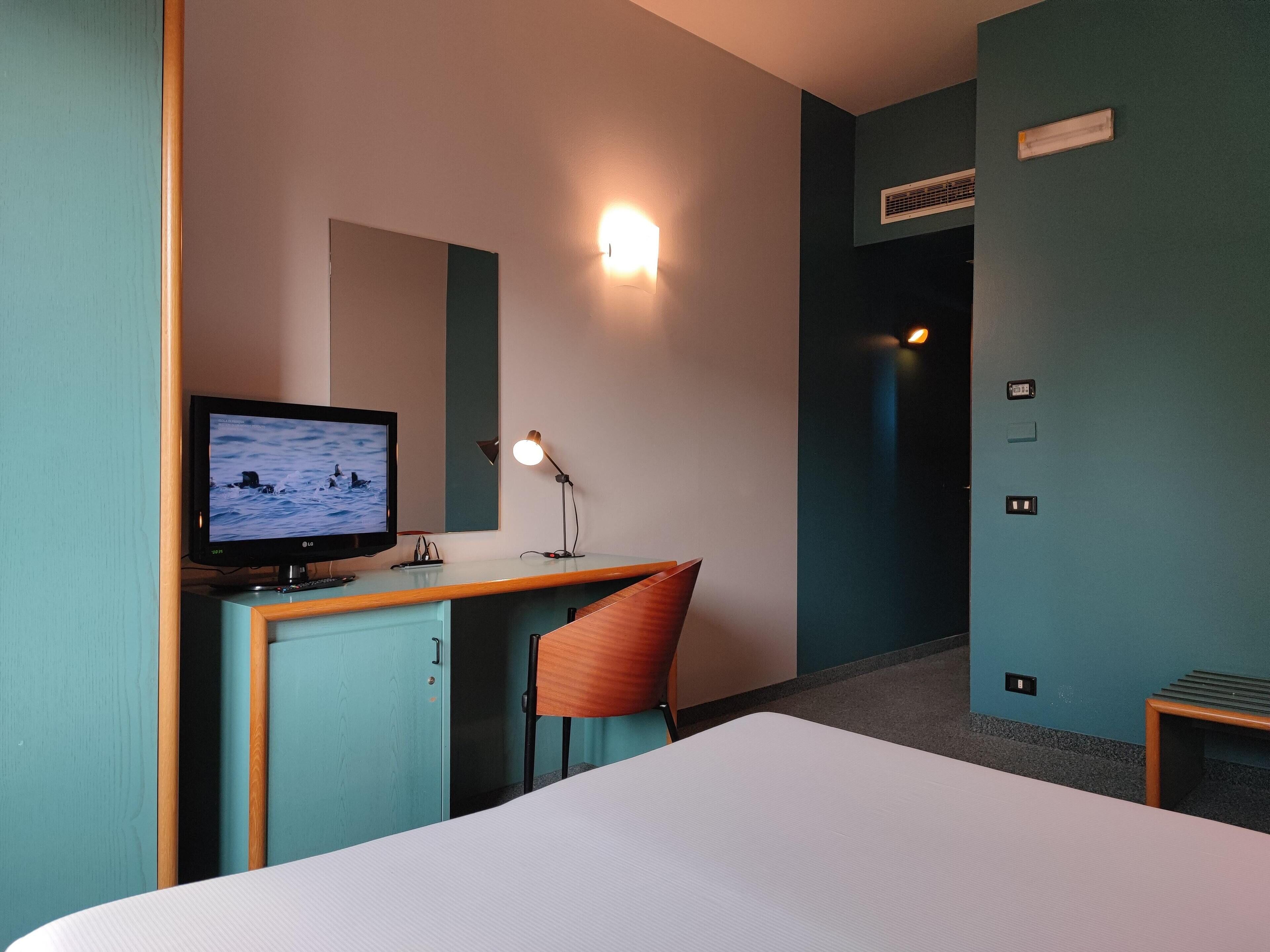 Una habitación de hotel con una cama, un escritorio con monitor de computadora, una silla y una lámpara fijada a la pared.