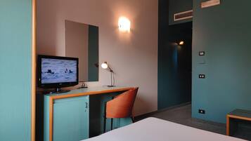 Una habitación de hotel con una cama, un escritorio con monitor de computadora, una silla y una lámpara fijada a la pared.