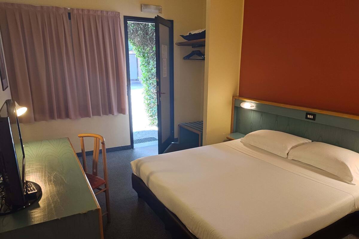 A hotel room with a bed, a desk with a chair, a lamp, and a window with curtains.