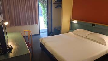 A hotel room with a bed, a desk with a chair, a lamp, and a window with curtains.